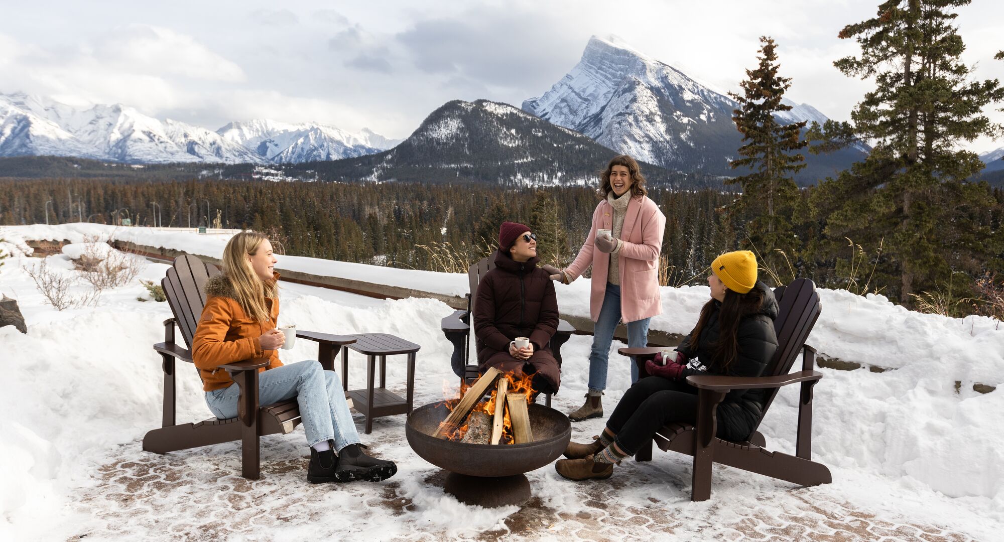 Friends enjoy warm drinks around the camp fire at Juniper Bistro with snowy Mount Rundle behind them in Banff National Park.